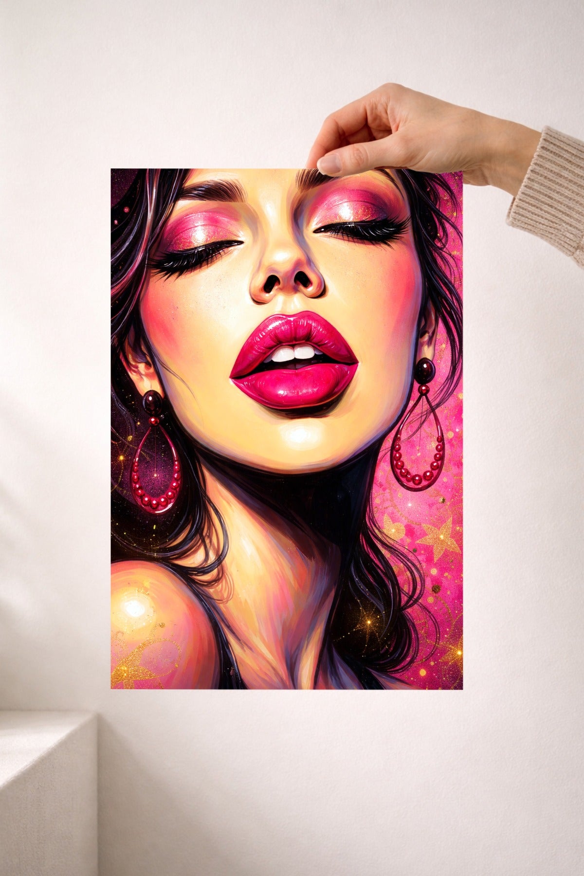 Hand hält ein Frauenportrait mit glänzenden pinken Lippen, rosa Glitzer-Augenmake-up, dunklen Haaren und dekorativen pinken Ohrringen vor einer weißen Wand.